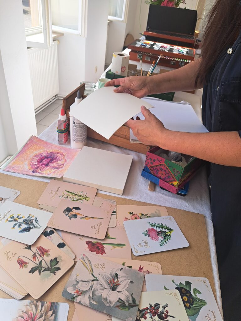 Botanische Karten mit Blumensymbolik und verschiedene Malmaterialien wie Acrylfarben, Ölkreiden und Pinseln auf einem großen Arbeitstisch im Kunsttreff freiRAUM.